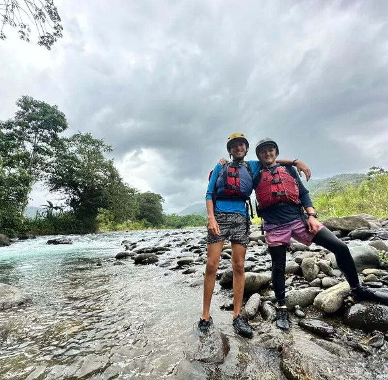 Two people wearing helmets and life jackets stand on rocks in a river. The water is flowing rapidly around them. They appear to be preparing for or have just finished a water activity like rafting or kayaking. The sky is overcast, and there are trees and vegetation on the riverbank.